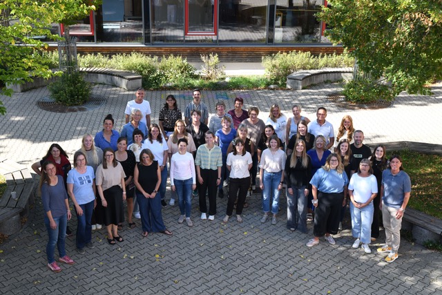 Das Foto zeigt die Kolleginnen und Kollegen der Eichenäcker-Schule. Sie stehen versammelt auf dem Pausenhof unter 2 Bäumen.